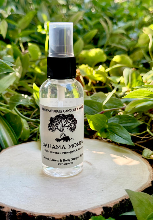 A clear two ounce finger-tip spray bottle with black and white label on a wooden surface with green leaves in the background. The bottle contains the scent "Bahama Momma".