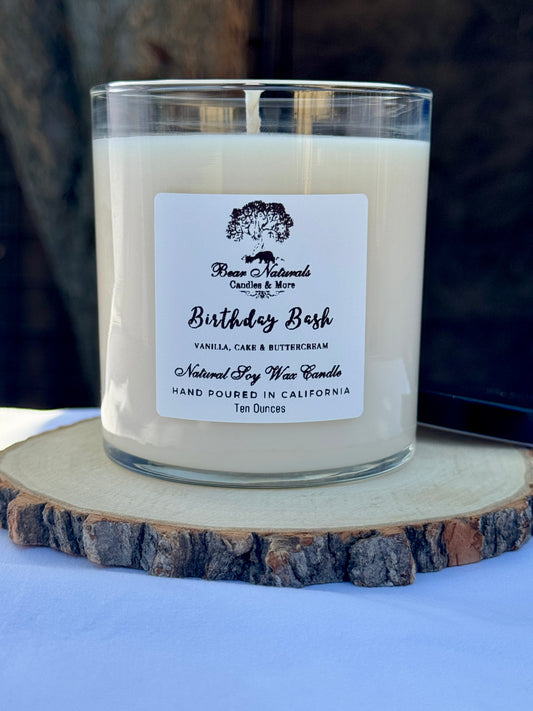 A ten ounce white candle in a clear glass jar with a black metal lid and white label with black writing sitting on a wooden surface. The candle is called "Birthday Bash".
