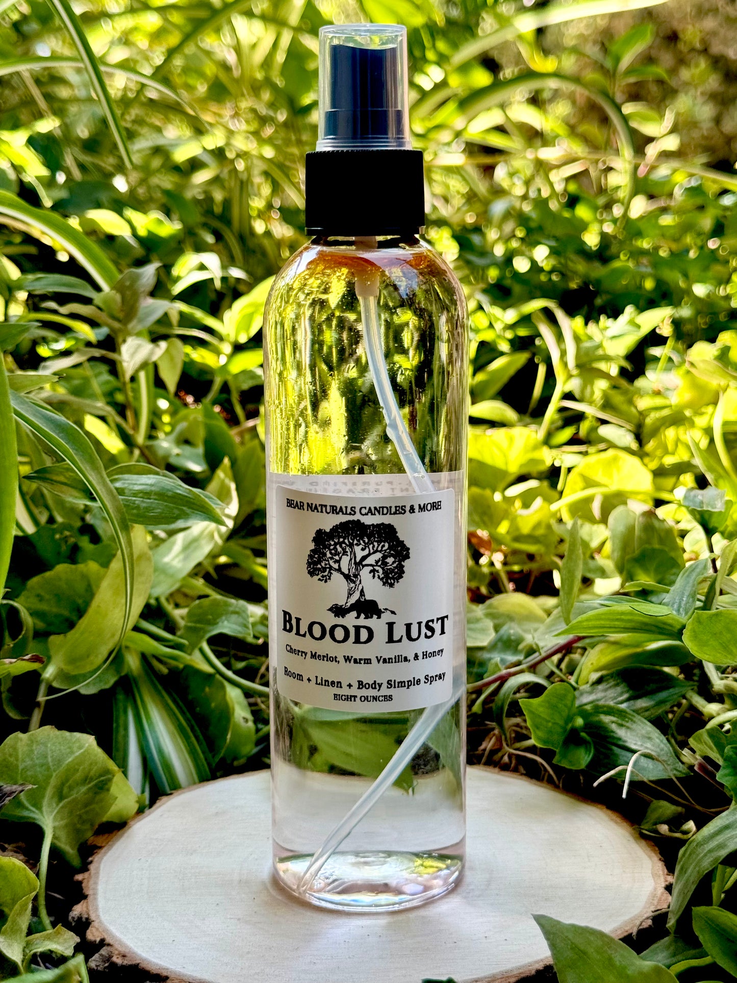A clear 8 ounce finger-tip spray bottle with black and white label on a wooden surface with green leaves in the background. The bottle contains the scent "Blood Lust".