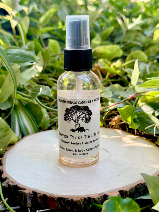 A clear two ounce finger-tip spray bottle with black and white label on a wooden surface with green leaves in the background. The bottle contains the scent "Driver Picks The Music".