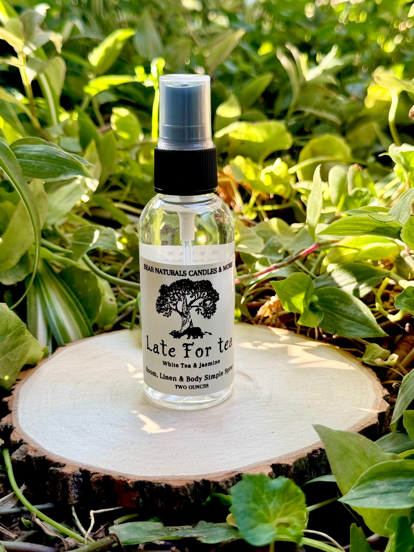 A clear two ounce finger-tip spray bottle with black and white label on a wooden surface with green leaves in the background. The bottle contains the scent "Late For Tea".