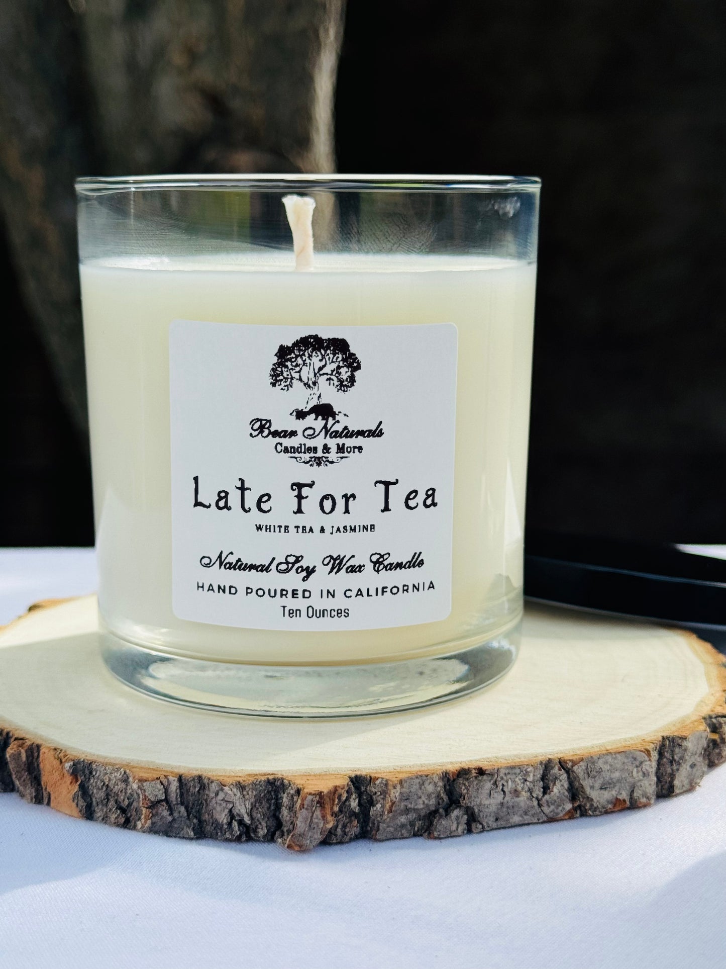 A white ten ounce candle in a clear glass jar with a black metal lid and white label with black writing sitting on a wooden surface. The candle is called "Late For Tea".