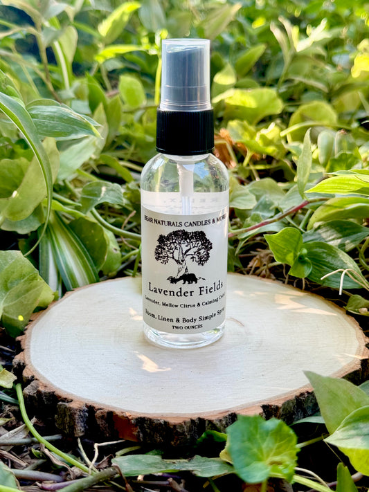 A clear two ounce finger-tip spray bottle with black and white label on a wooden surface with green leaves in the background. The bottle contains the scent "Lavender Fields".
