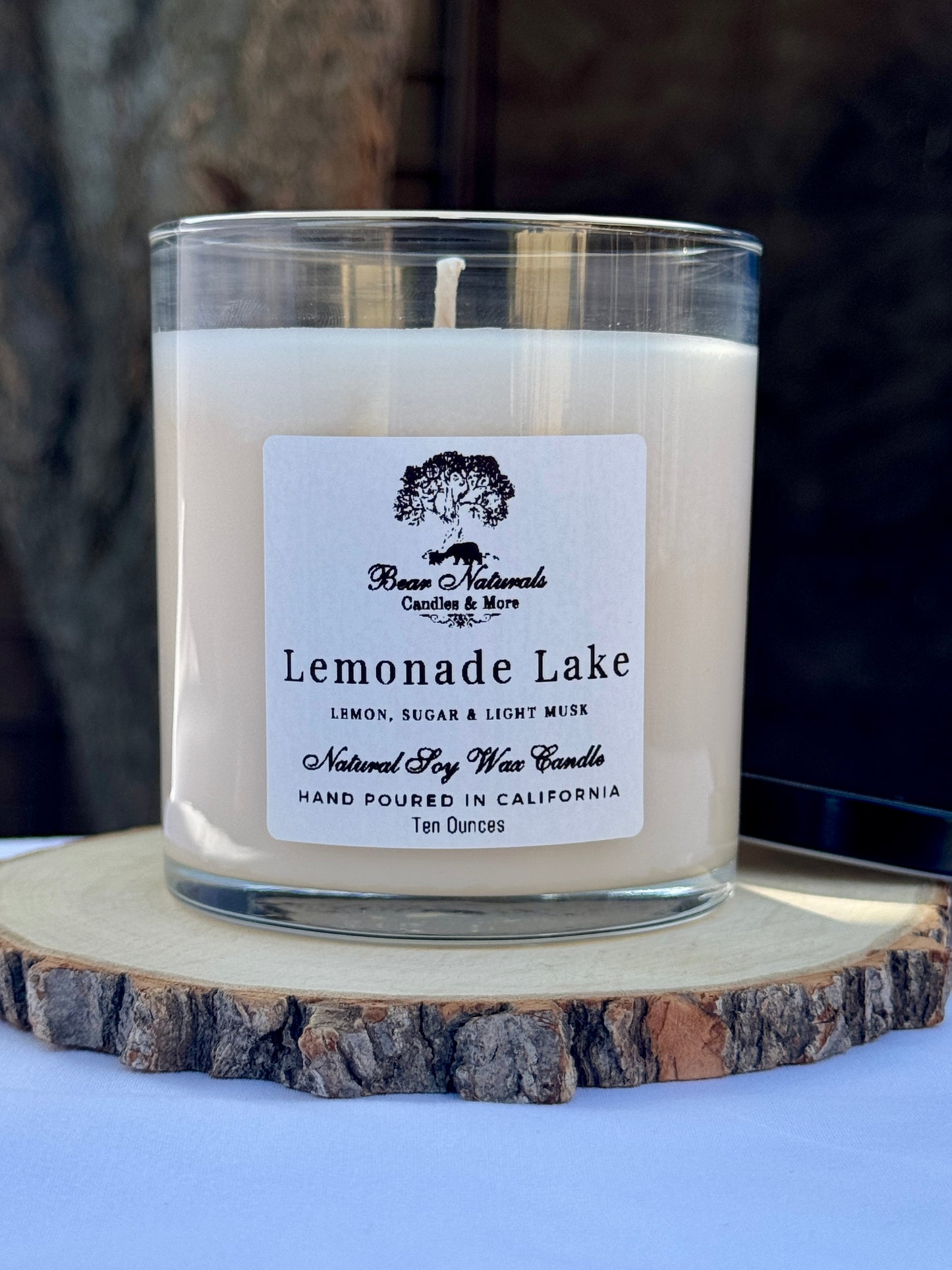 A white ten ounce candle in a clear glass jar with a black metal lid and white label with black writing sitting on a wooden surface. The candle is called "Lemonade Lake".