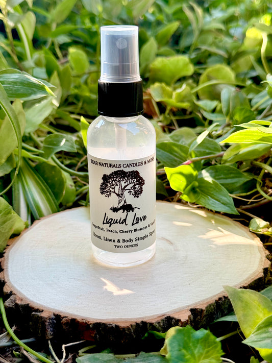 A clear two ounce finger-tip spray bottle with black and white label on a wooden surface with green leaves in the background. The bottle contains the scent "Liquid Love".
