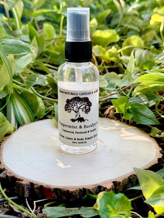 A clear two ounce finger-tip spray bottle with black and white label on a wooden surface with green leaves in the background. The bottle contains the scent "Peppermint & Eucalyptus".