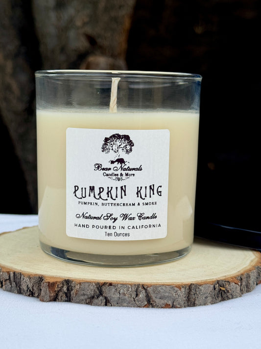 A white ten ounce candle in a clear glass jar with a black metal lid and white label with black writing sitting on a wooden surface. The candle is called "Pumpkin King".