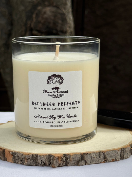 A white ten ounce candle in a clear glass jar with a black metal lid and white label with black writing sitting on a wooden surface. The candle is called "Reindeer Presents".