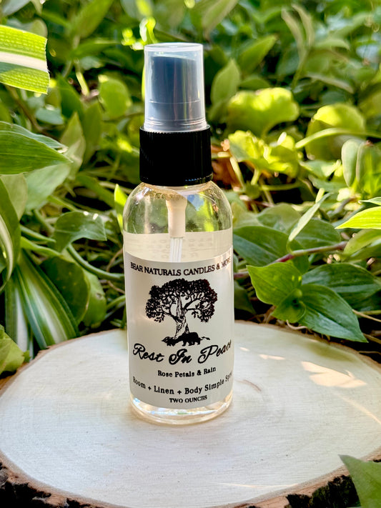 A clear two ounce finger-tip spray bottle with black and white label on a wooden surface with green leaves in the background. The bottle contains the scent "Rest In Peace".