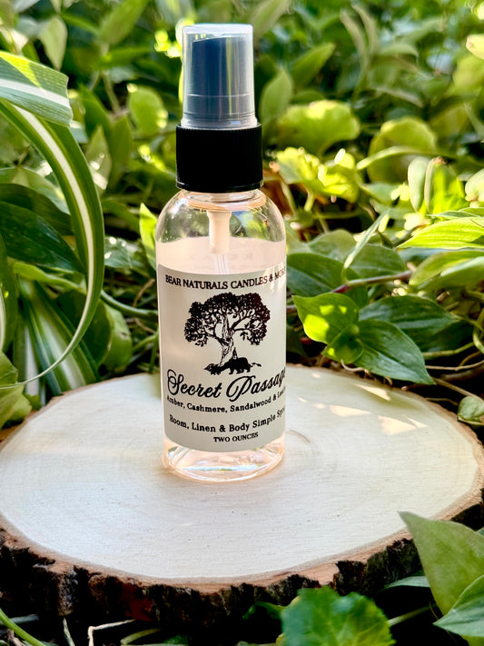 A clear two ounce finger-tip spray bottle with black and white label on a wooden surface with green leaves in the background. The bottle contains the scent "Secret Passage".