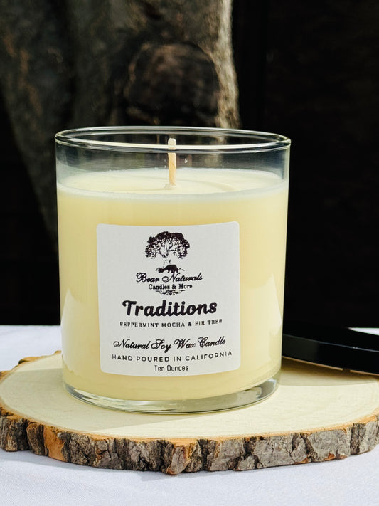 A white ten ounce candle in a clear glass jar with a black metal lid and white label with black writing sitting on a wooden surface. The candle is called "Traditions".