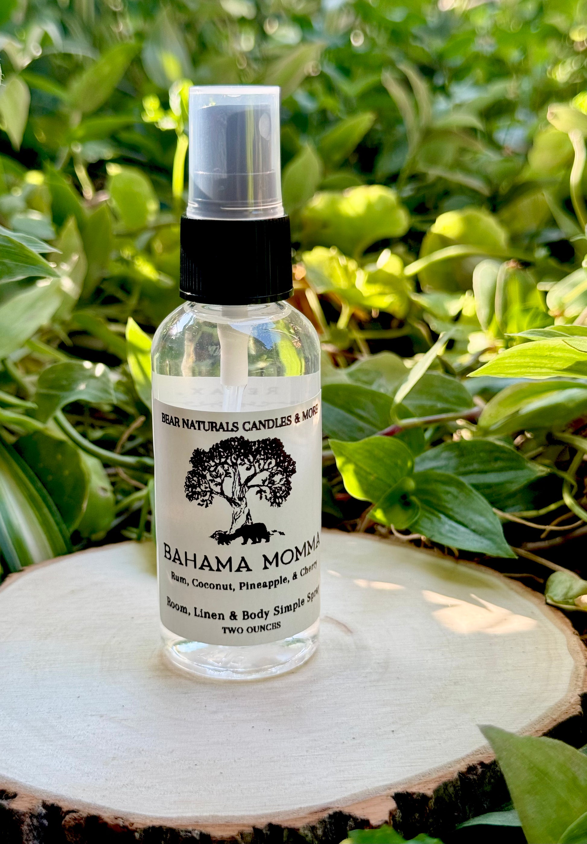 A clear two ounce finger-tip spray bottle with black and white label on a wooden surface with green leaves in the background. The bottle contains the scent "Bahama Momma".