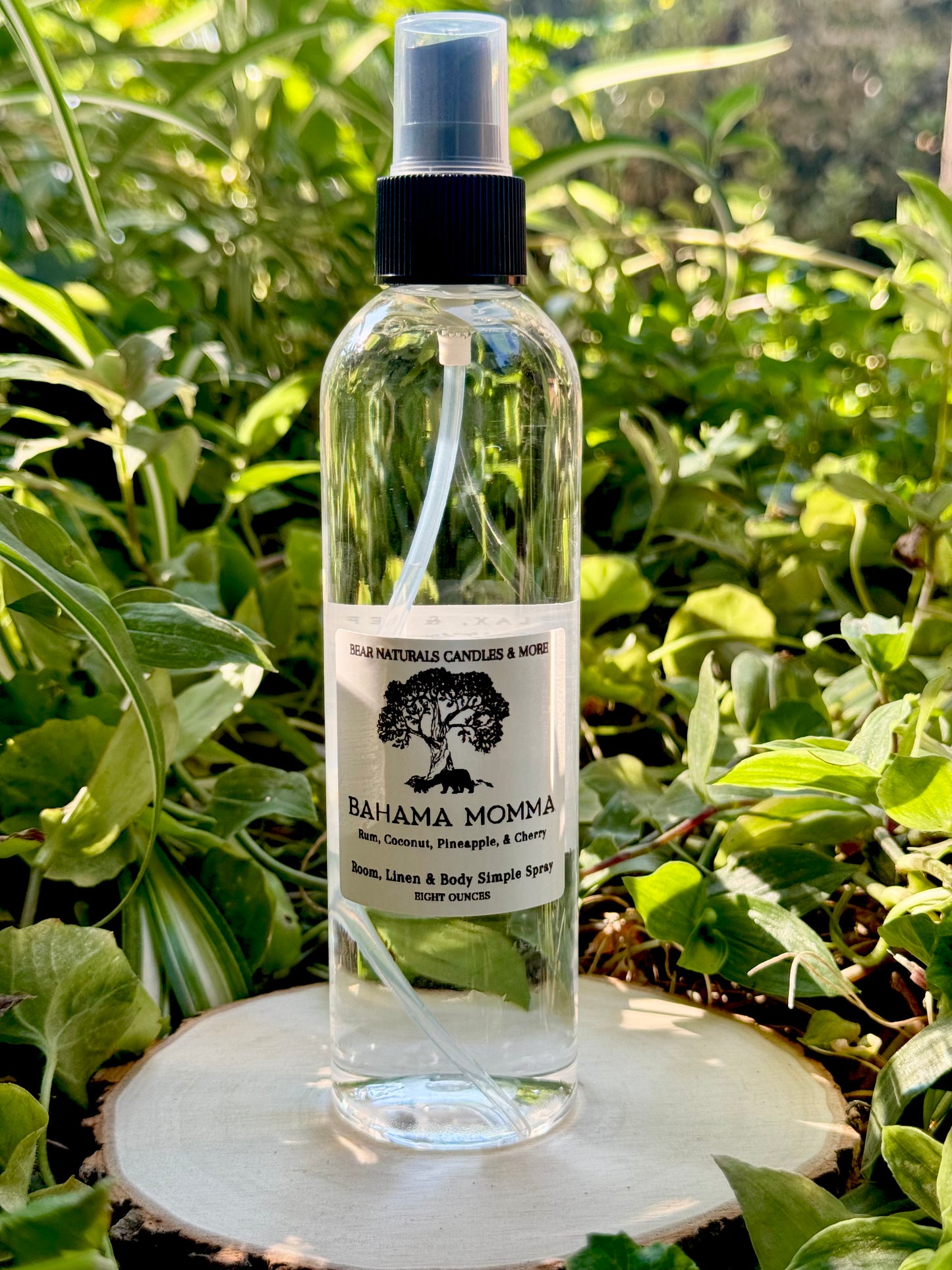 A clear eight ounce finger-tip spray bottle with black and white label on a wooden surface with green leaves in the background. The bottle contains the scent "Bahama Momma".