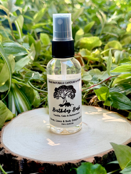 A Clear two ounce finger-tip spray bottle with black and white label on a wooden surface with green leaves in the background. The bottle contains the scent "Birthday Bash".

