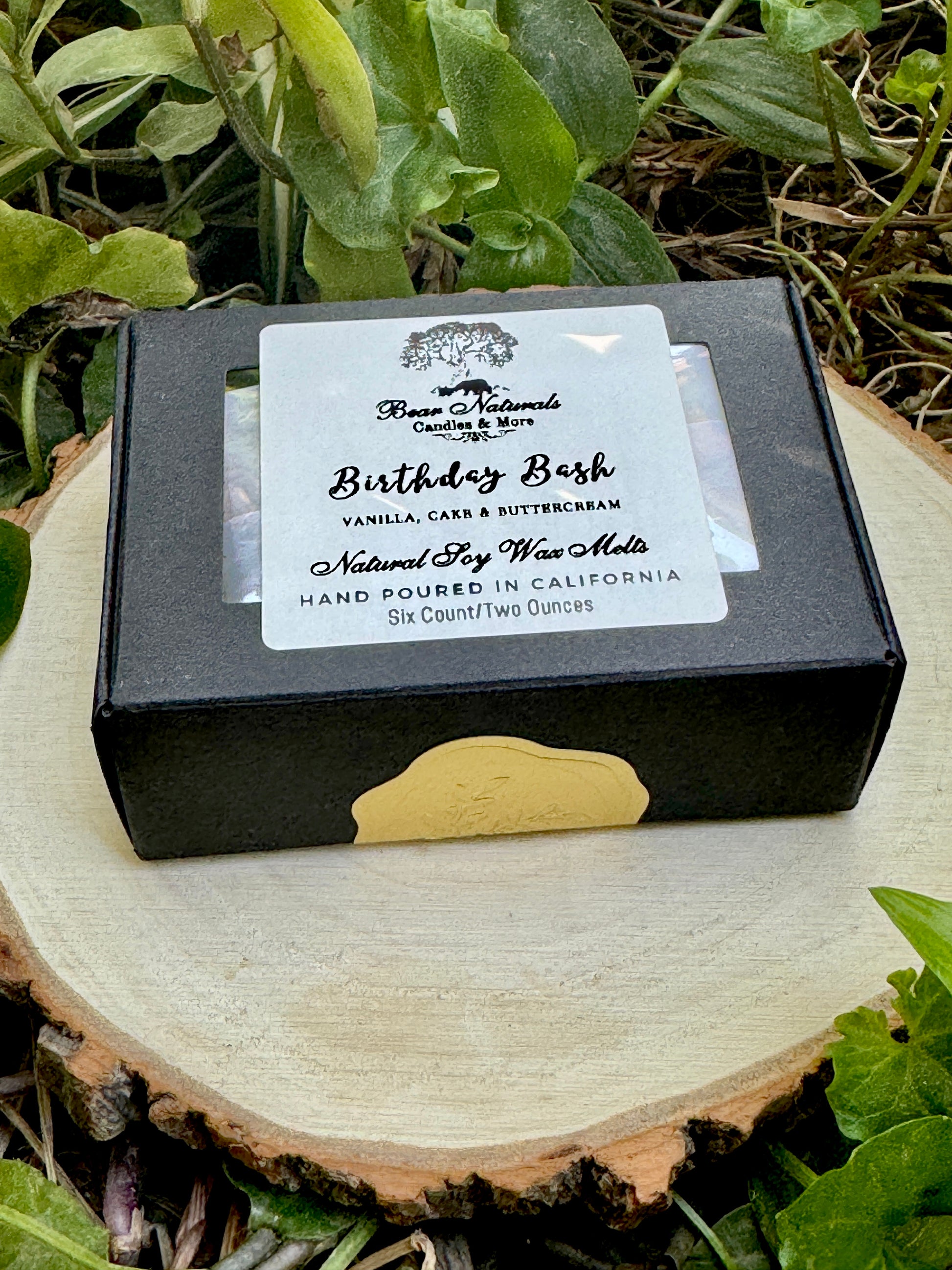 A small, hand-sized black box with a white label featuring black writing that says "Birthday Bash" sealed with a small gold sticker sitting on a wooden surface with green leaves in the background. 