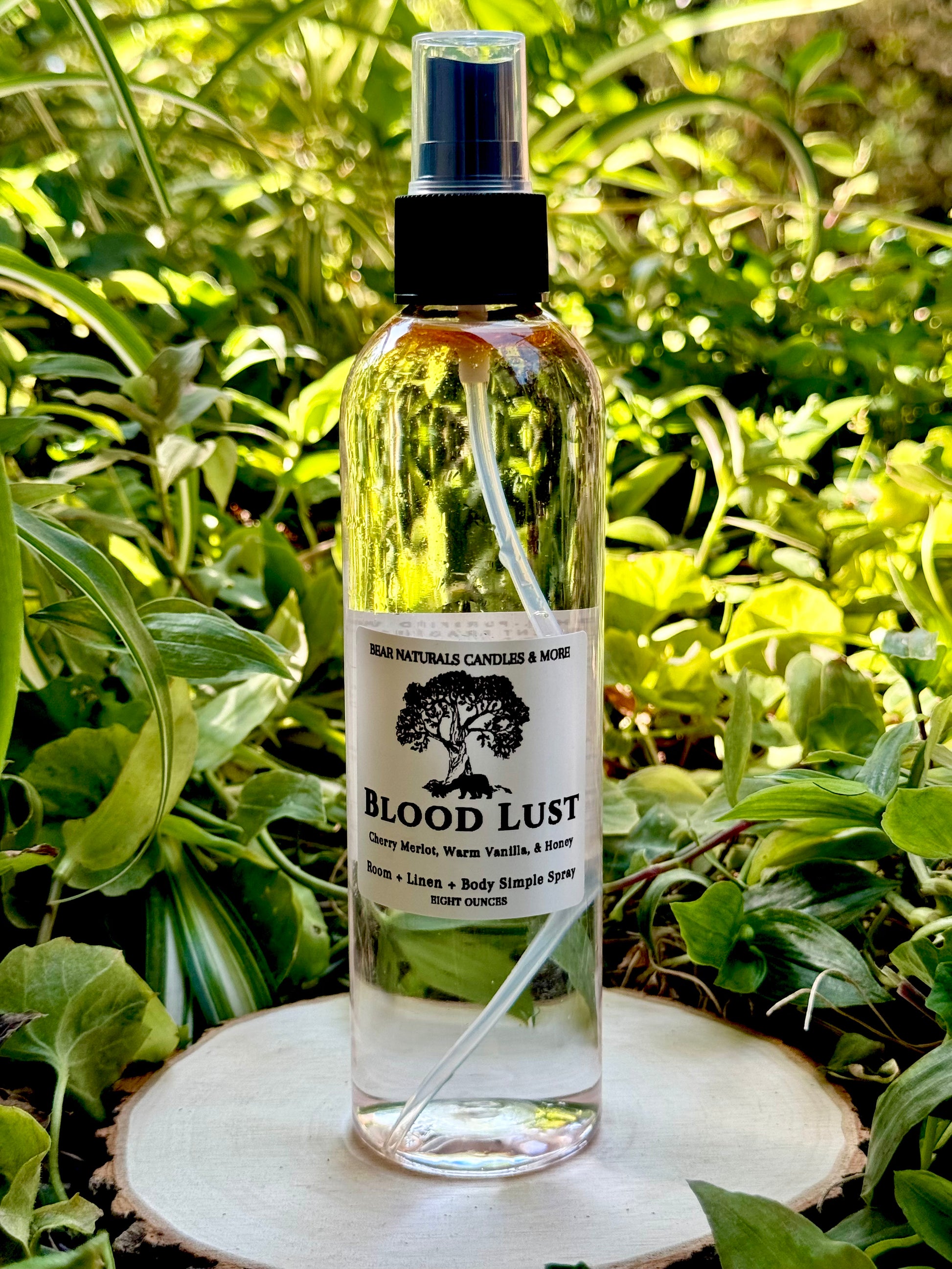 A clear 8 ounce finger-tip spray bottle with black and white label on a wooden surface with green leaves in the background. The bottle contains the scent "Blood Lust".