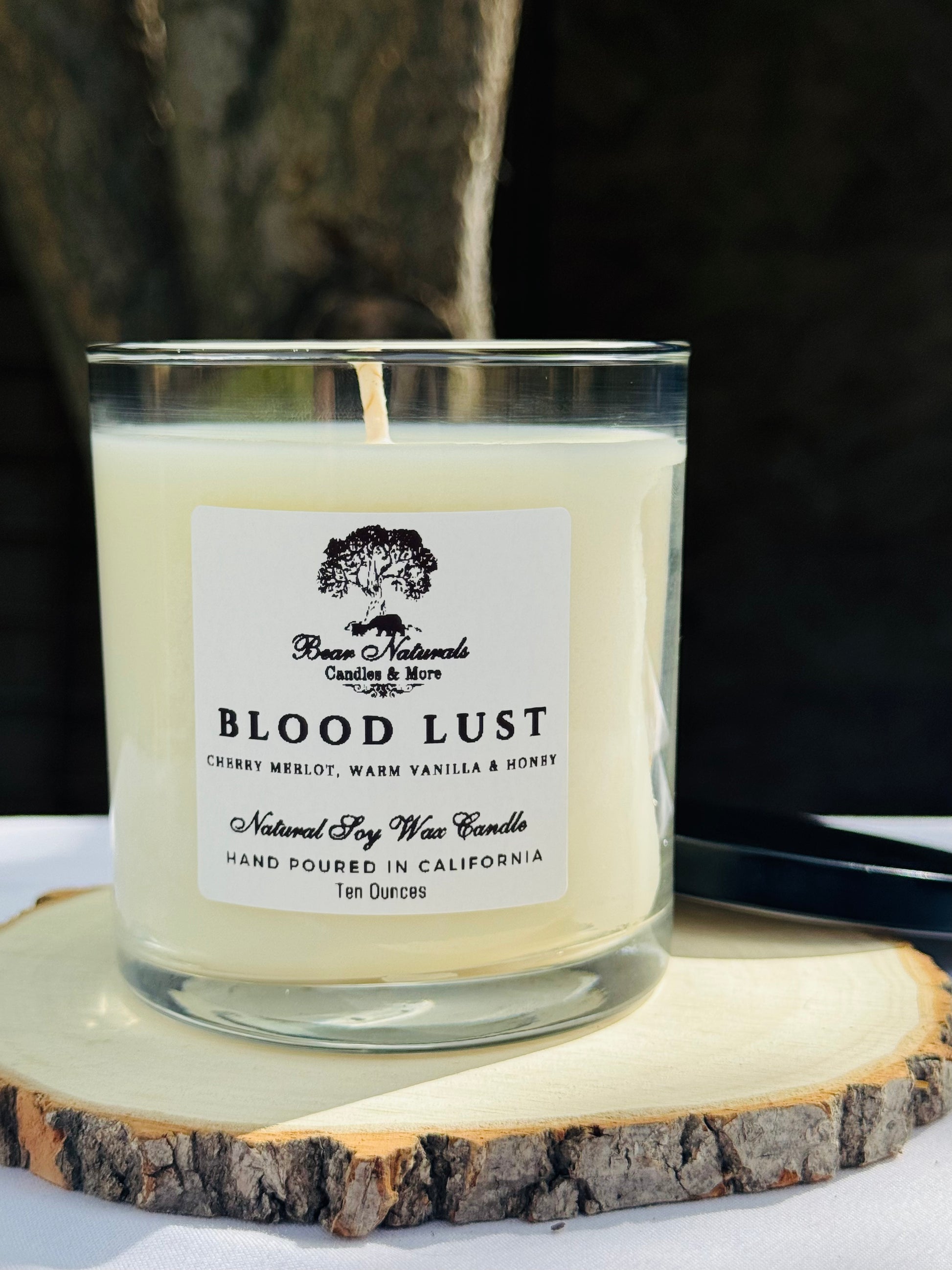 A ten ounce white scented candle in a clear glass jar with a black metal lid and white label with black writing sitting on a wooden surface. The candle is called "Blood Lust".
