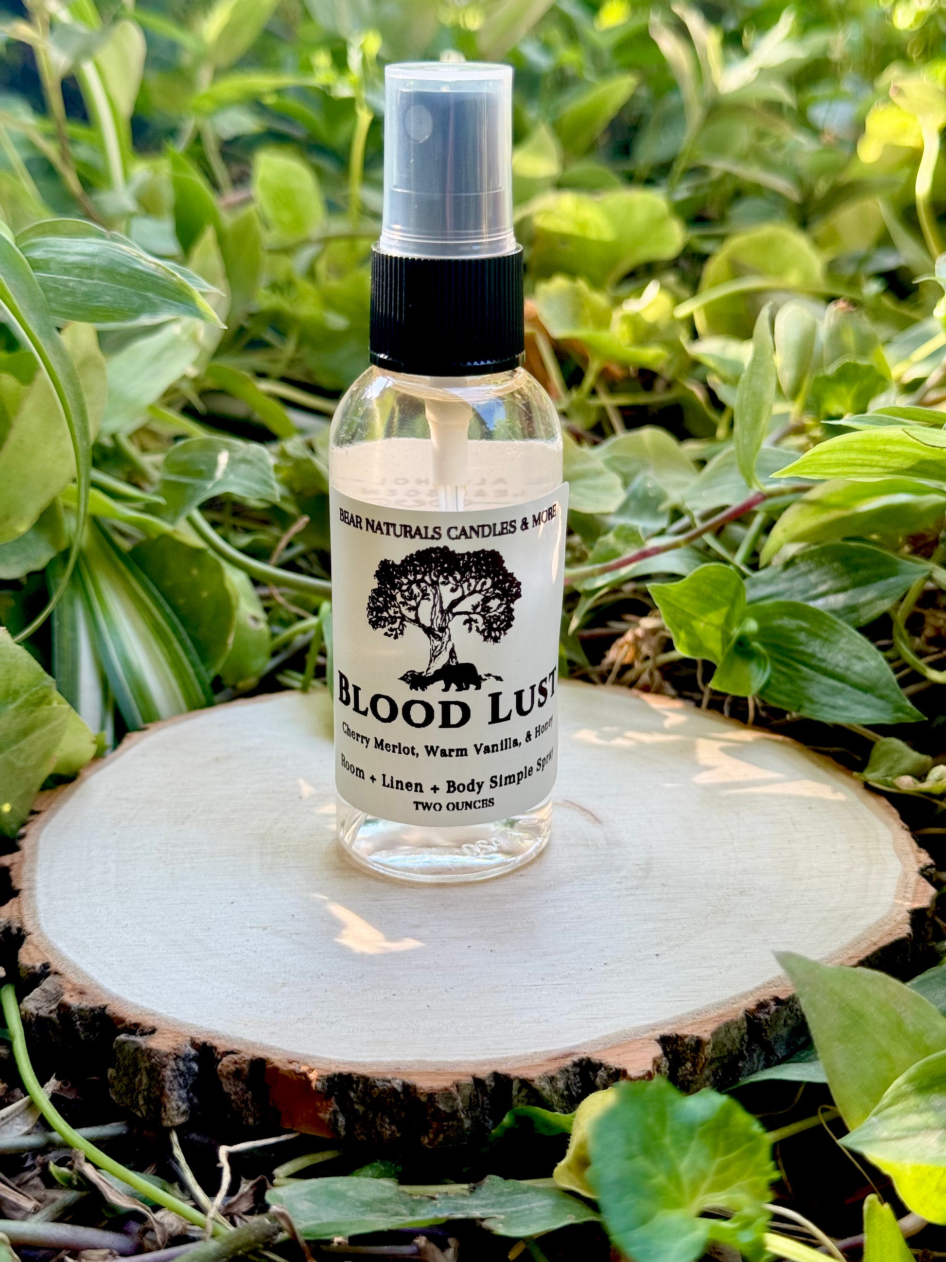 A clear 2 ounce finger-tip spray bottle with black and white label on a wooden surface with green leaves in the background. The bottle contains the scent "Blood Lust".