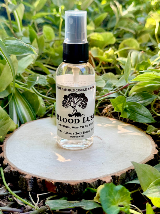 A clear 2 ounce finger-tip spray bottle with black and white label on a wooden surface with green leaves in the background. The bottle contains the scent "Blood Lust".