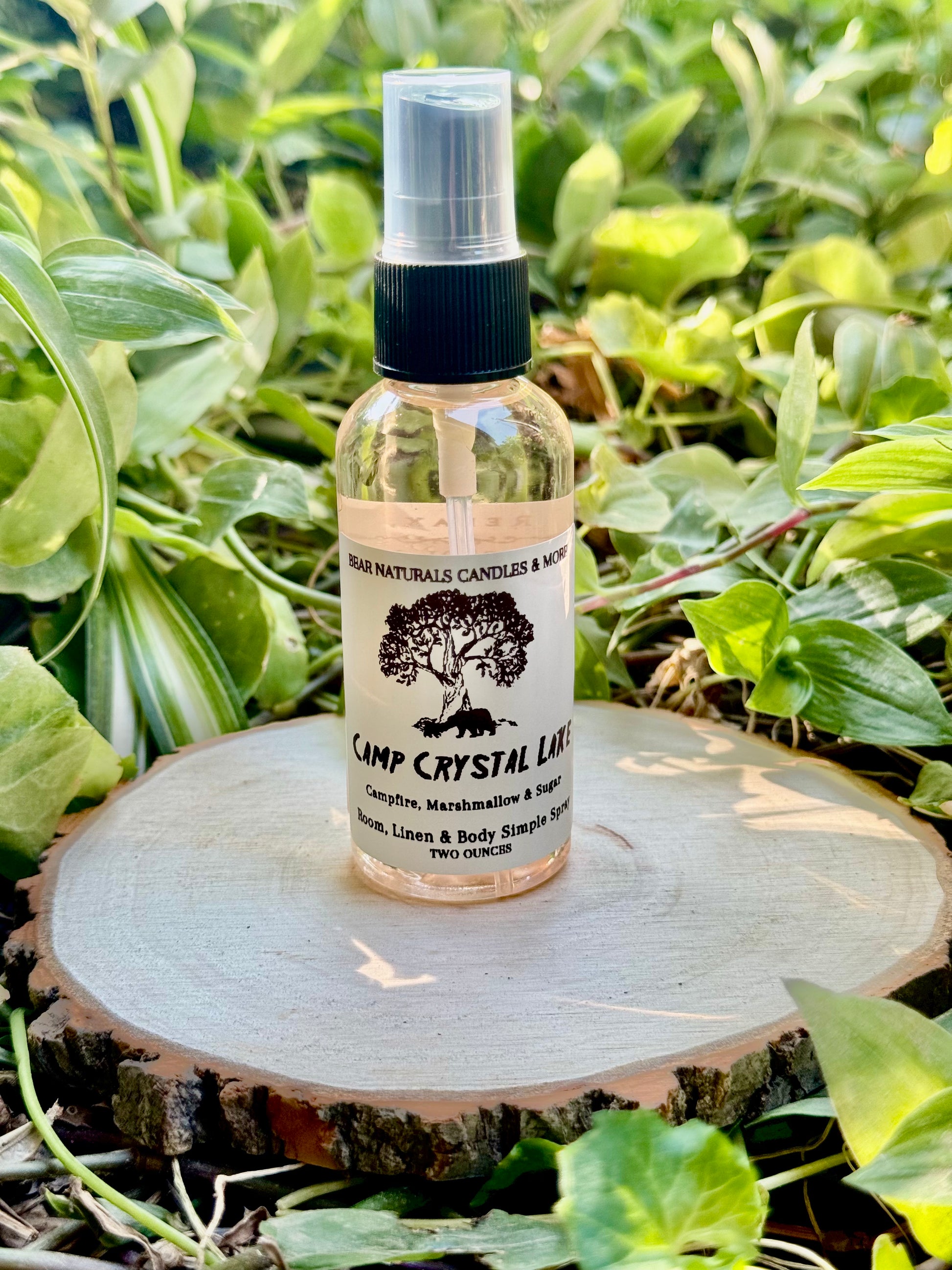 Clear finger-tip spray bottle with black and white label on a wooden surface with green leaves in the background. The bottle contains the scent "Camp Crystal Lake".
