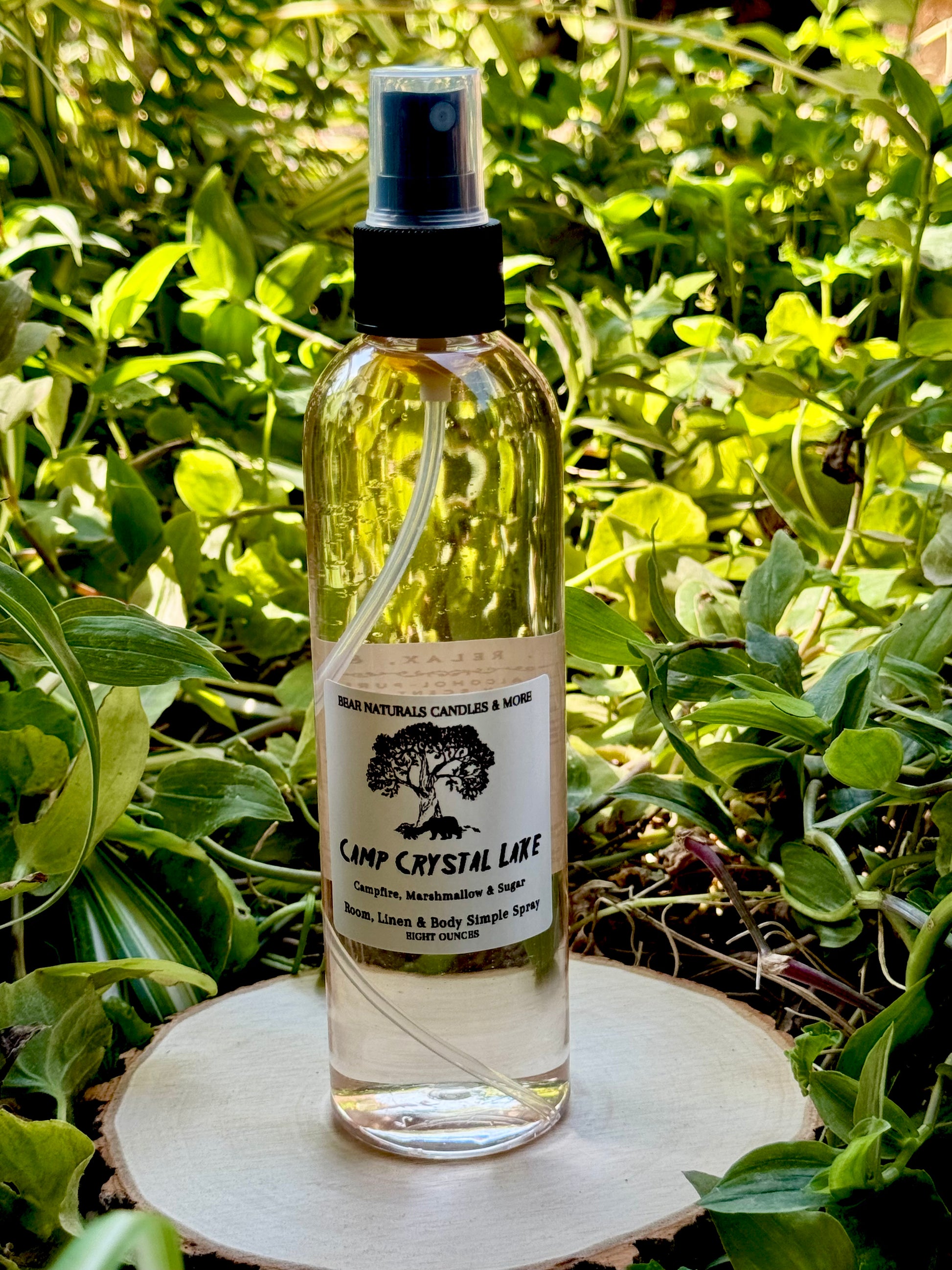 Clear finger-tip spray bottle with black and white label on a wooden surface with green leaves in the background. The bottle contains the scent "Camp Crystal Lake".