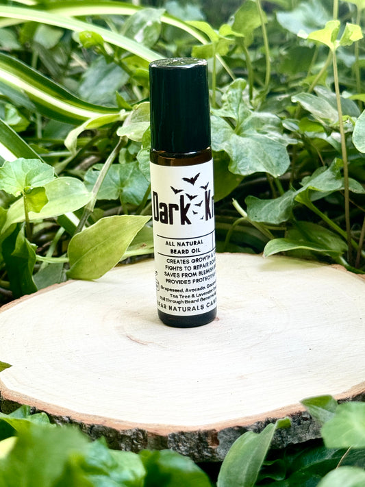 10ml amber colored roll on bottle with a black lid featuring a white label reading "Dark Knight All Natural Beard Oil" sitting on a wooden surface with green leaves in the background