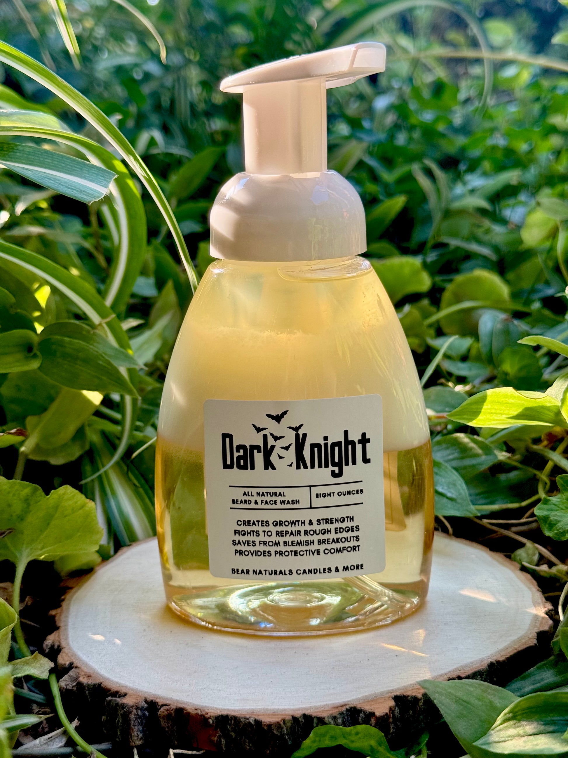 an 8oz clear bottle with a white foaming pump lid featuring a white label with black writing that reads "Dark Knight All Natural Foaming Beard And Face Wash" sitting on a wooden surface with green leaves in the background.