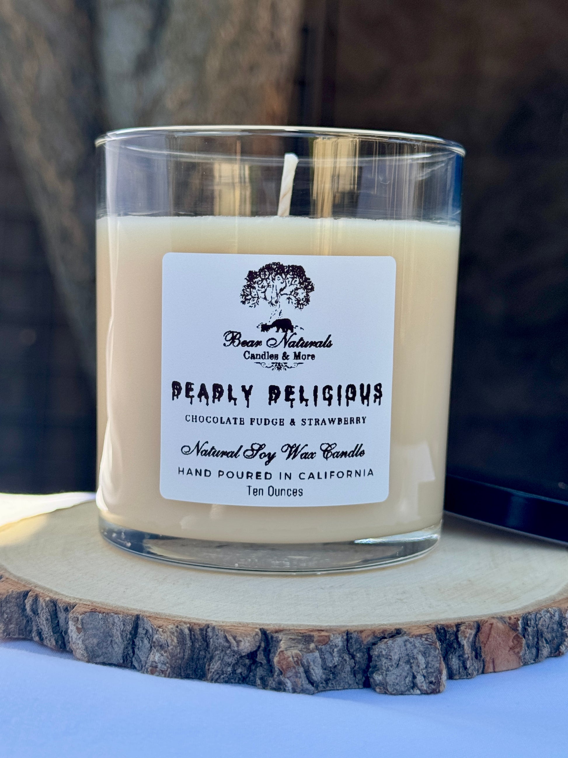 A ten ounce white scented candle in a clear glass jar with a black metal lid and white label with black writing sitting on a wooden surface. The candle is called "Deadly Delicious". 