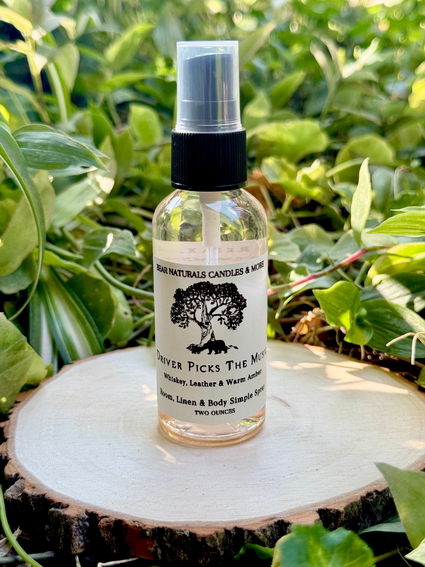 A clear two ounce finger-tip spray bottle with black and white label on a wooden surface with green leaves in the background. The bottle contains the scent "Driver Picks The Music".