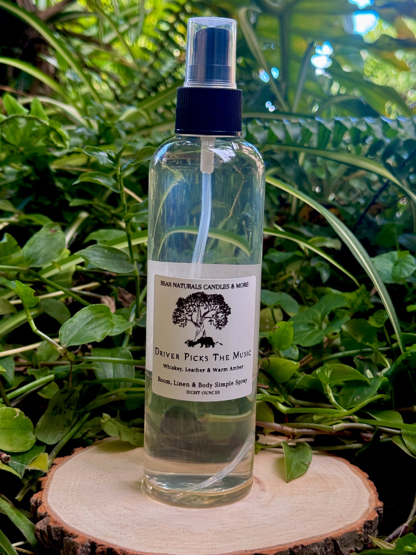 A clear eight ounce finger-tip spray bottle with black and white label on a wooden surface with green leaves in the background. The bottle contains the scent "Driver Picks The Music".