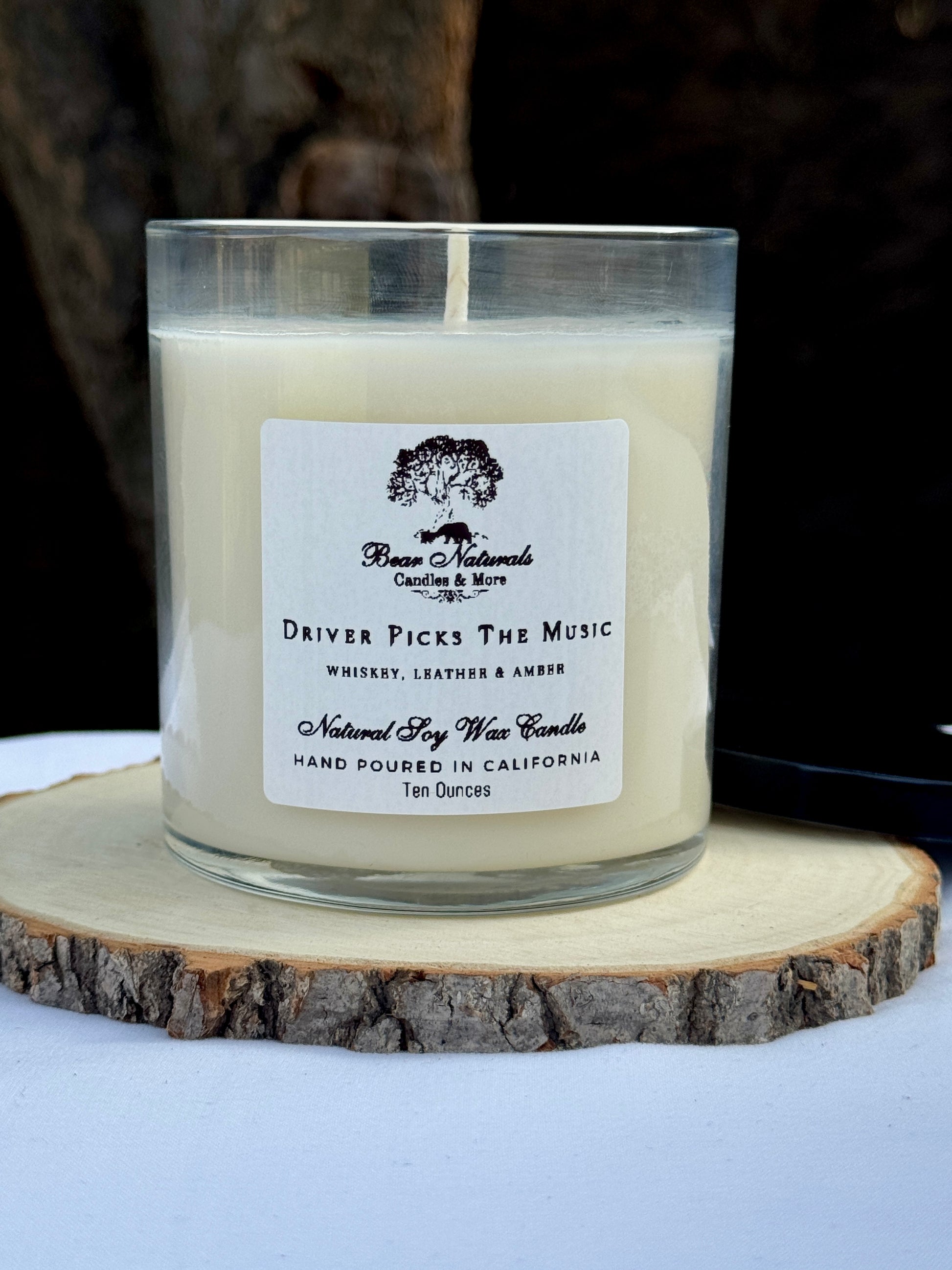 A white ten ounce candle in a clear glass jar with a black metal lid and white label with black writing sitting on a wooden surface. The candle is called "Driver Picks The Music".