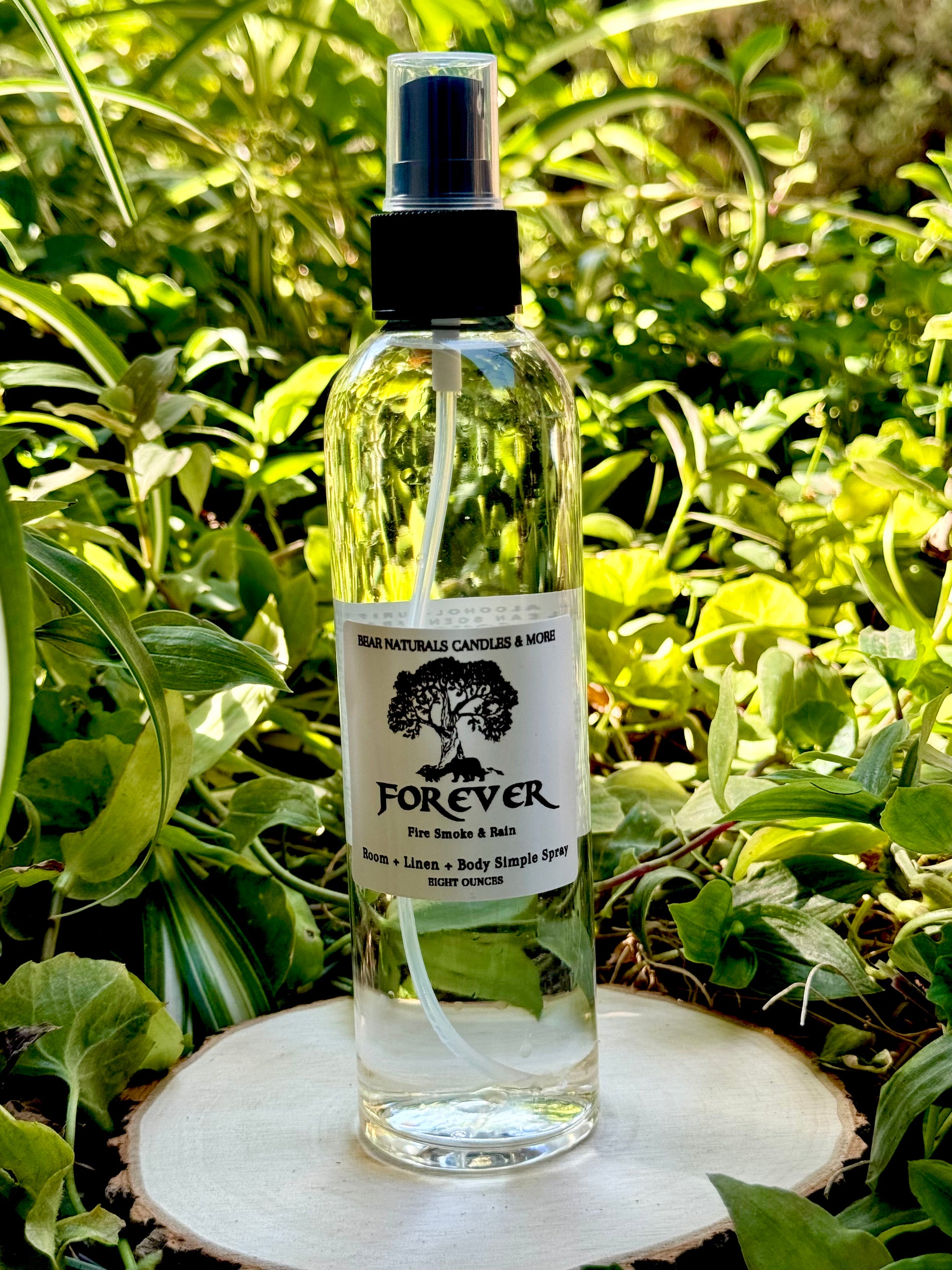 A clear eight ounce finger-tip spray bottle with black and white label on a wooden surface with green leaves in the background. The bottle contains the scent "Forever".