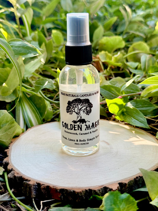 A clear two ounce finger-tip spray bottle with black and white label on a wooden surface with green leaves in the background. The bottle contains the scent "Golden Magic".