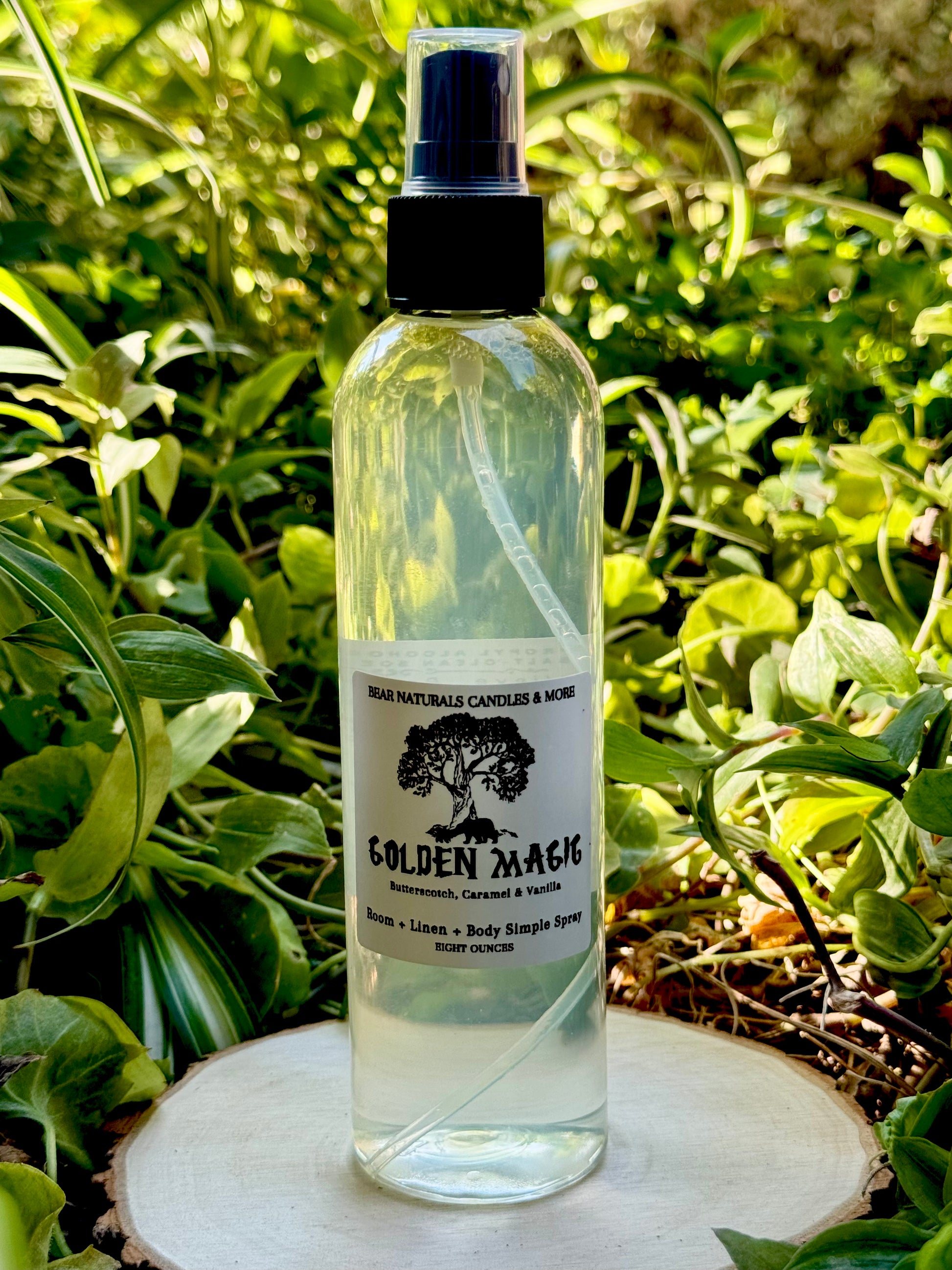 A clear eight ounce finger-tip spray bottle with black and white label on a wooden surface with green leaves in the background. The bottle contains the scent "Golden Magic".
