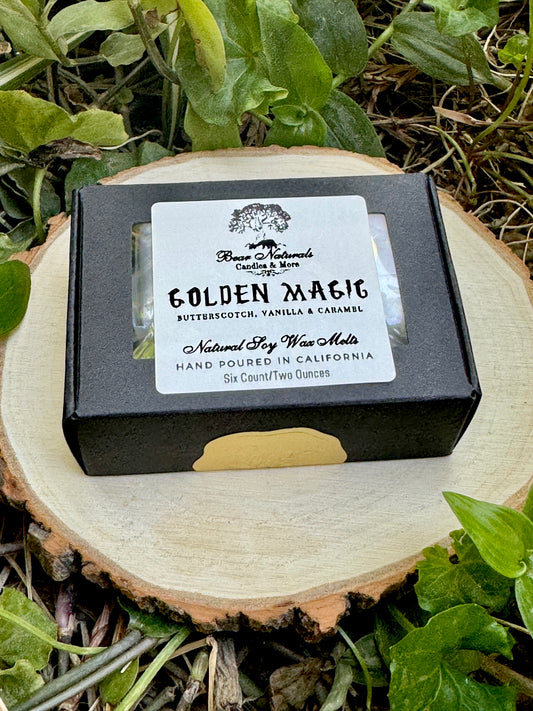 A small, hand-sized black box with a white label featuring black writing that says "Golden Magic" sealed with a small gold sticker sitting on a wooden surface with green leaves in the background.