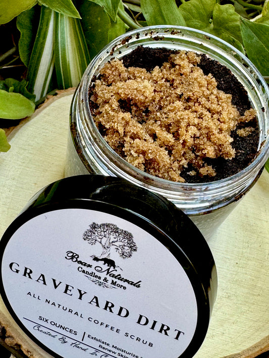 A six ounce clear jar containing exfoliating coffee scrub topped with brown sugar sitting on a wooden surface with green leaves in the background. The white label with black writing is featured on the lid and reads "Graveyard Dirt All Natural Coffee Scrub".