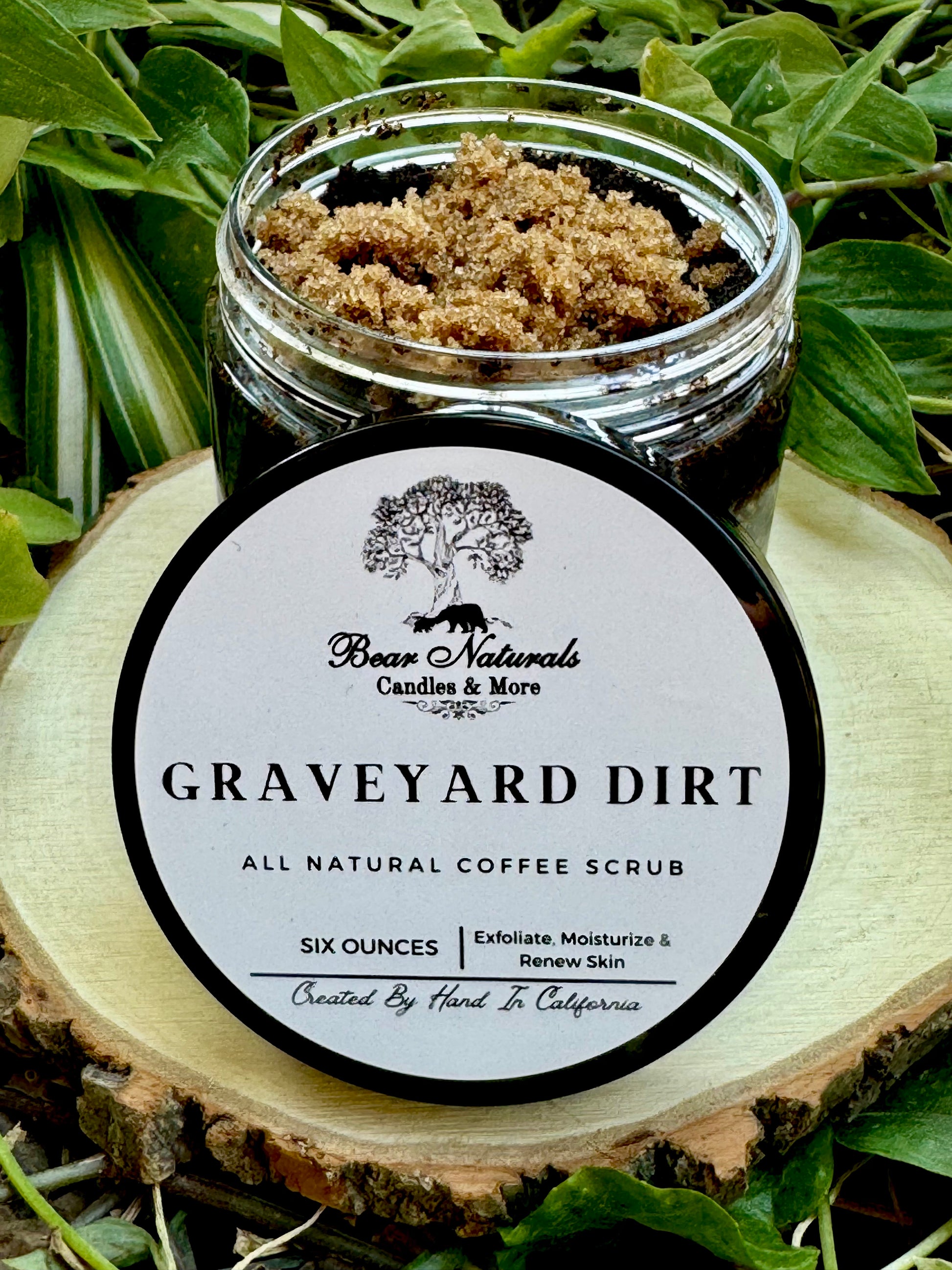A six ounce clear jar containing exfoliating coffee scrub topped with brown sugar sitting on a wooden surface with green leaves in the background. The white label with black writing is featured on the lid and reads "Graveyard Dirt All Natural Coffee Scrub".