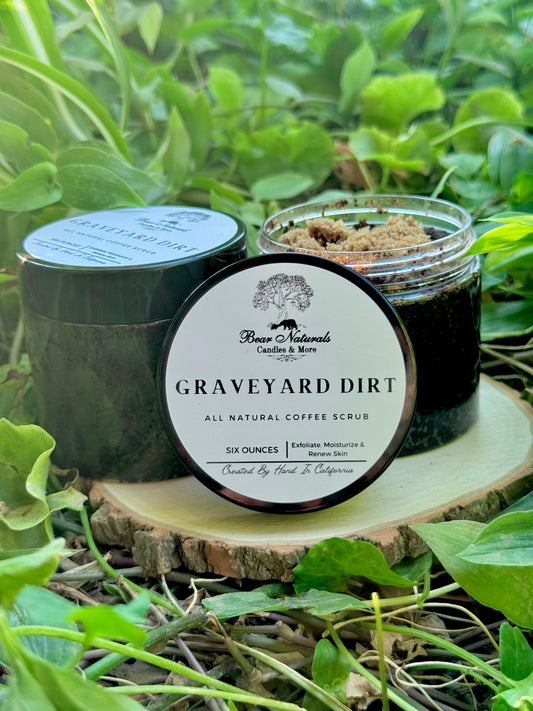 A six ounce clear jar containing exfoliating coffee scrub topped with brown sugar sitting on a wooden surface with green leaves in the background. The white label with black writing is featured on the lid and reads "Graveyard Dirt All Natural Coffee Scrub".