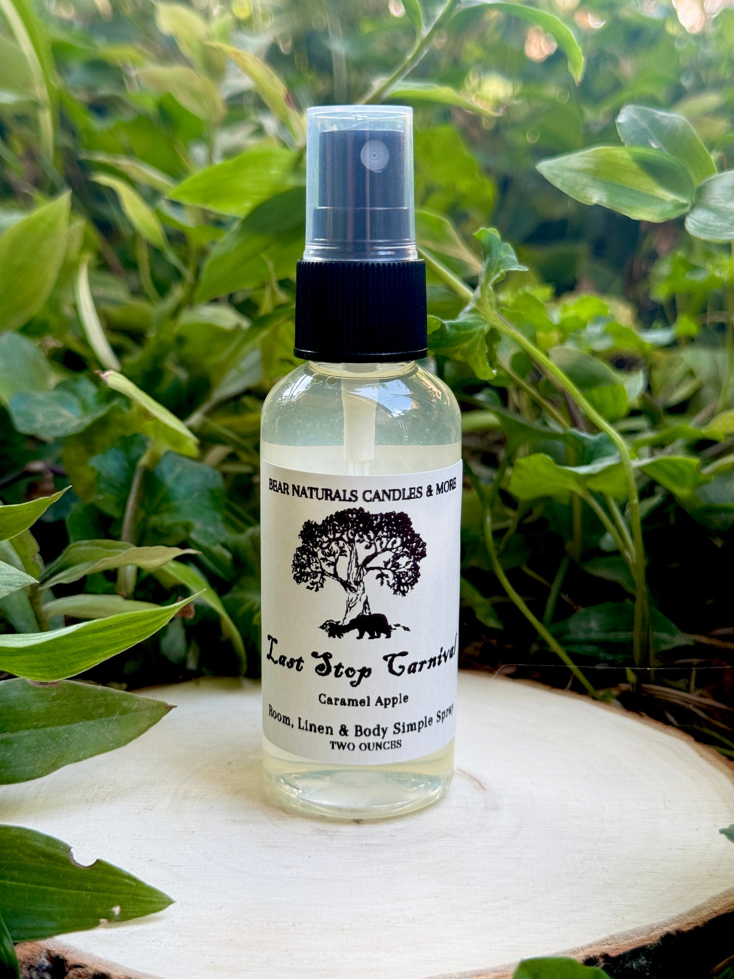 A clear two ounce finger-tip spray bottle with black and white label on a wooden surface with green leaves in the background. The bottle contains the scent "Last Stop Carnival".