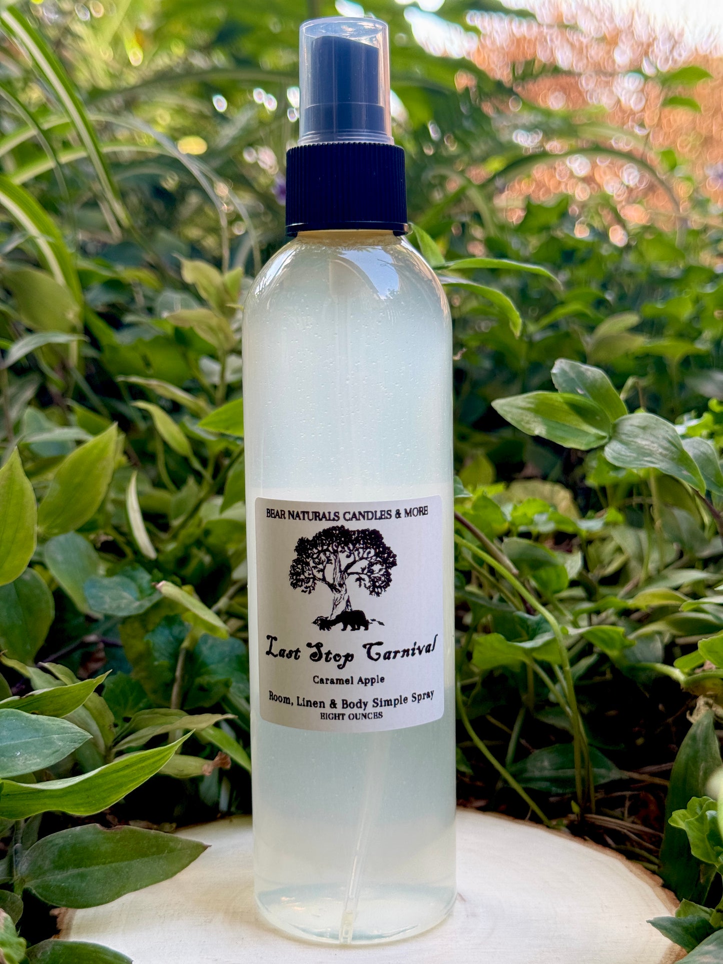 A clear eight ounce finger-tip spray bottle with black and white label on a wooden surface with green leaves in the background. The bottle contains the scent "Last Stop Carnival".