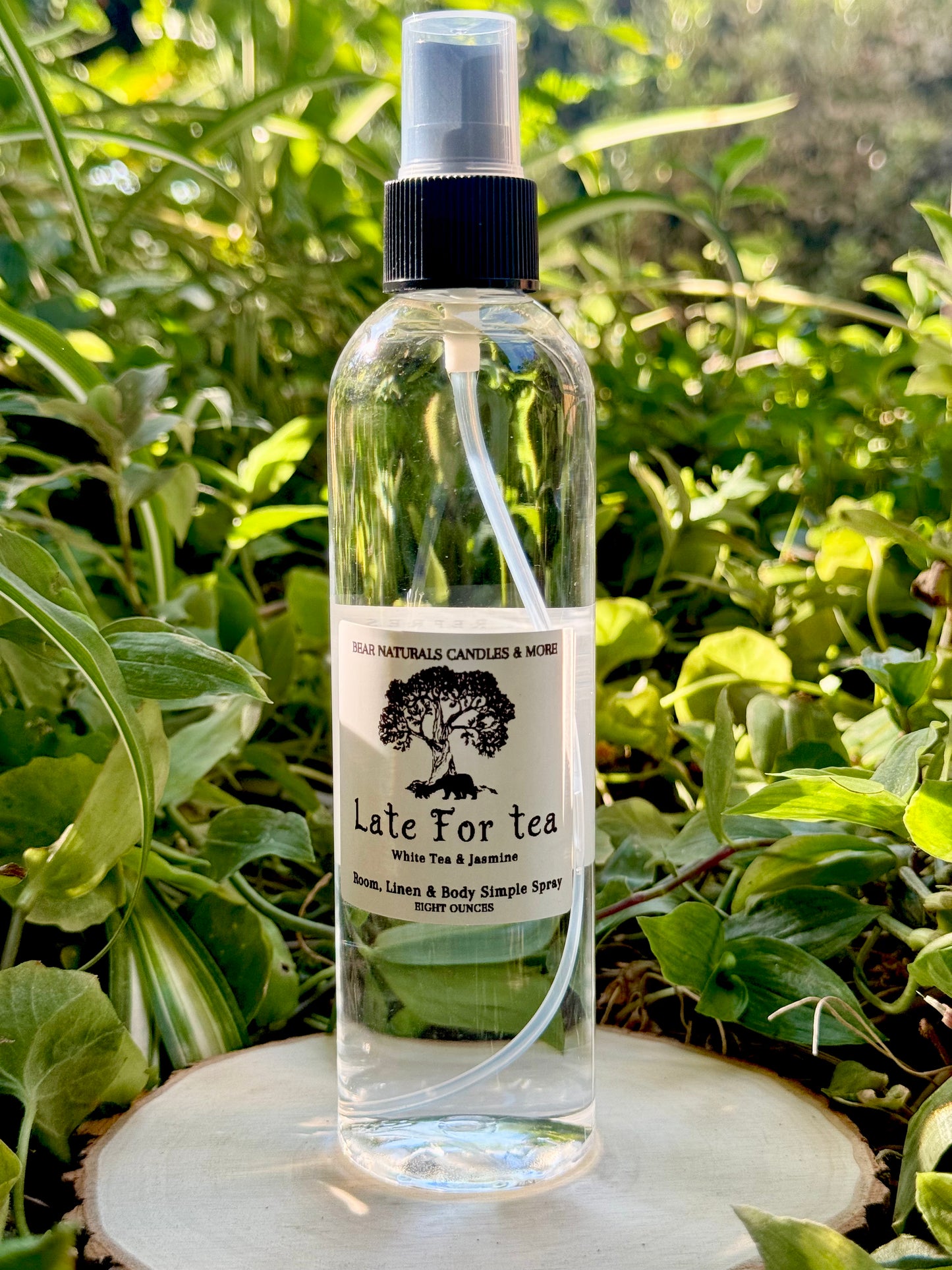 A clear eight ounce finger-tip spray bottle with black and white label on a wooden surface with green leaves in the background. The bottle contains the scent "Late For Tea".