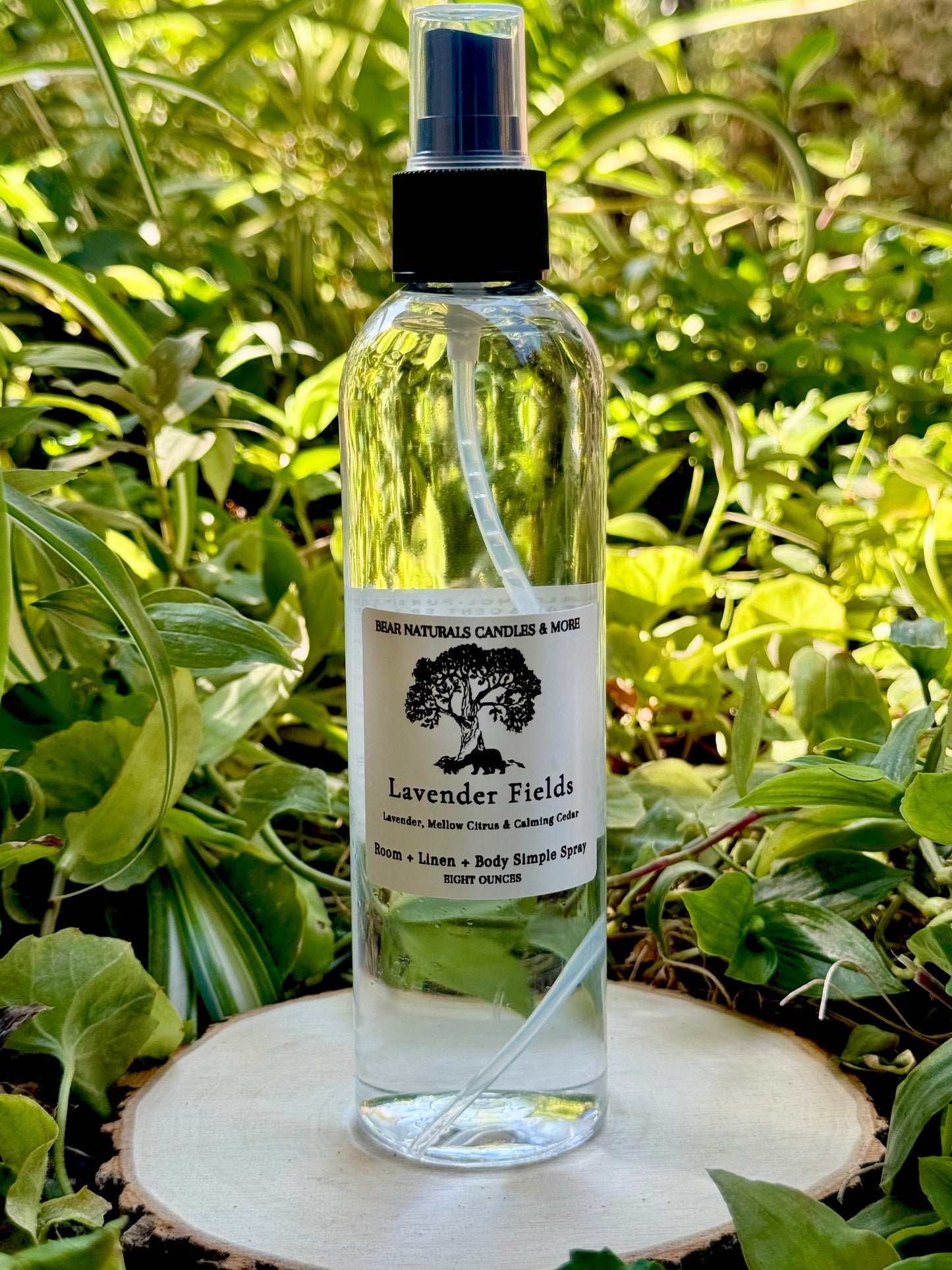A clear eight ounce finger-tip spray bottle with black and white label on a wooden surface with green leaves in the background. The bottle contains the scent "Lavender Fields".