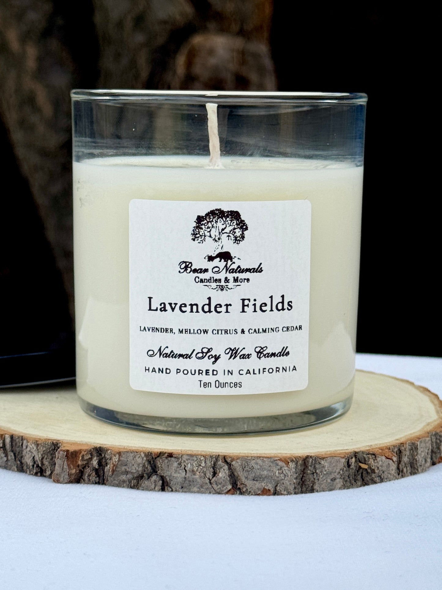 A white ten ounce candle in a clear glass jar with a black metal lid and white label with black writing sitting on a wooden surface. The candle is called "Lavender Fields".