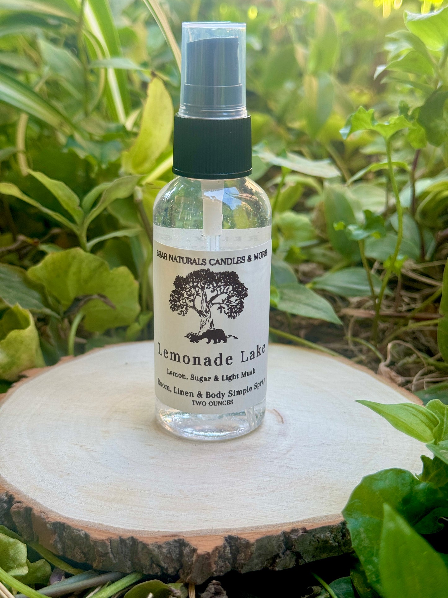 A clear two ounce finger-tip spray bottle with black and white label on a wooden surface with green leaves in the background. The bottle contains the scent "Lemonade Lake".

