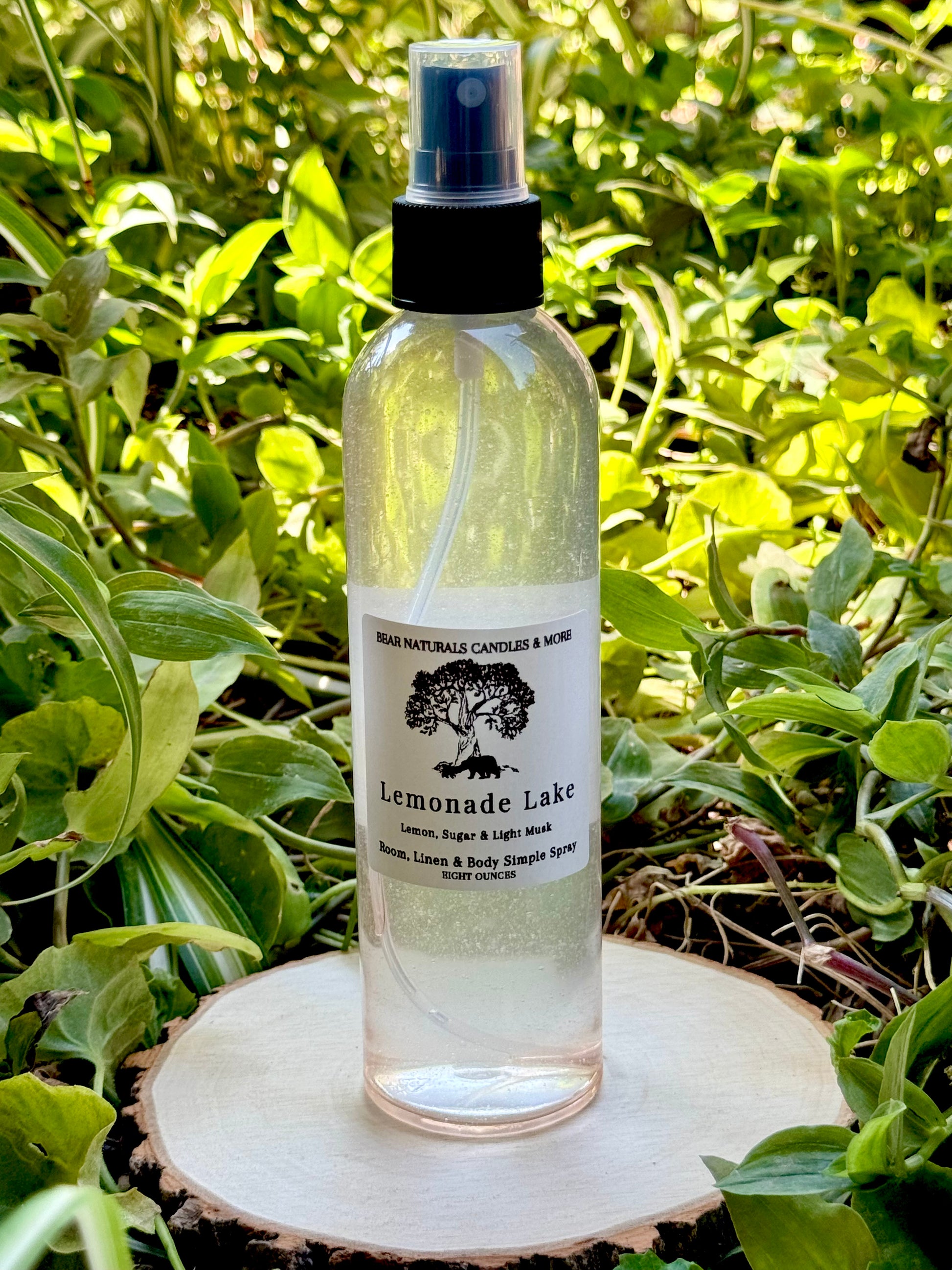 A clear eight ounce finger-tip spray bottle with black and white label on a wooden surface with green leaves in the background. The bottle contains the scent "Lemonade Lake".