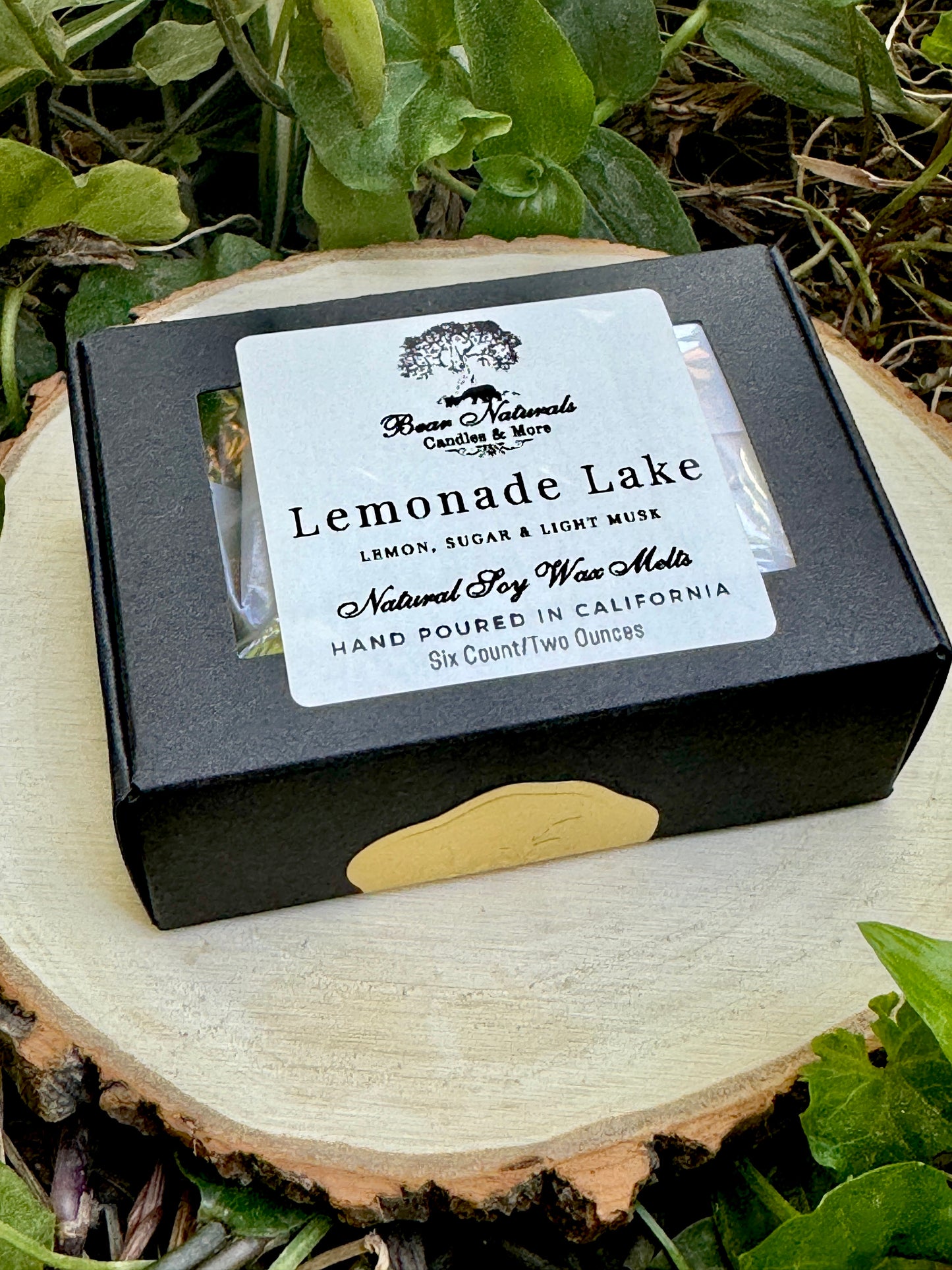 A small, hand-sized black box with a white label featuring black writing that says "Lemonade Lake" sealed with a small gold sticker sitting on a wooden surface with green leaves in the background. 