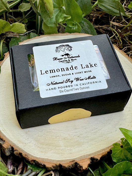 A small, hand-sized black box with a white label featuring black writing that says "Lemonade Lake" sealed with a small gold sticker sitting on a wooden surface with green leaves in the background. 
