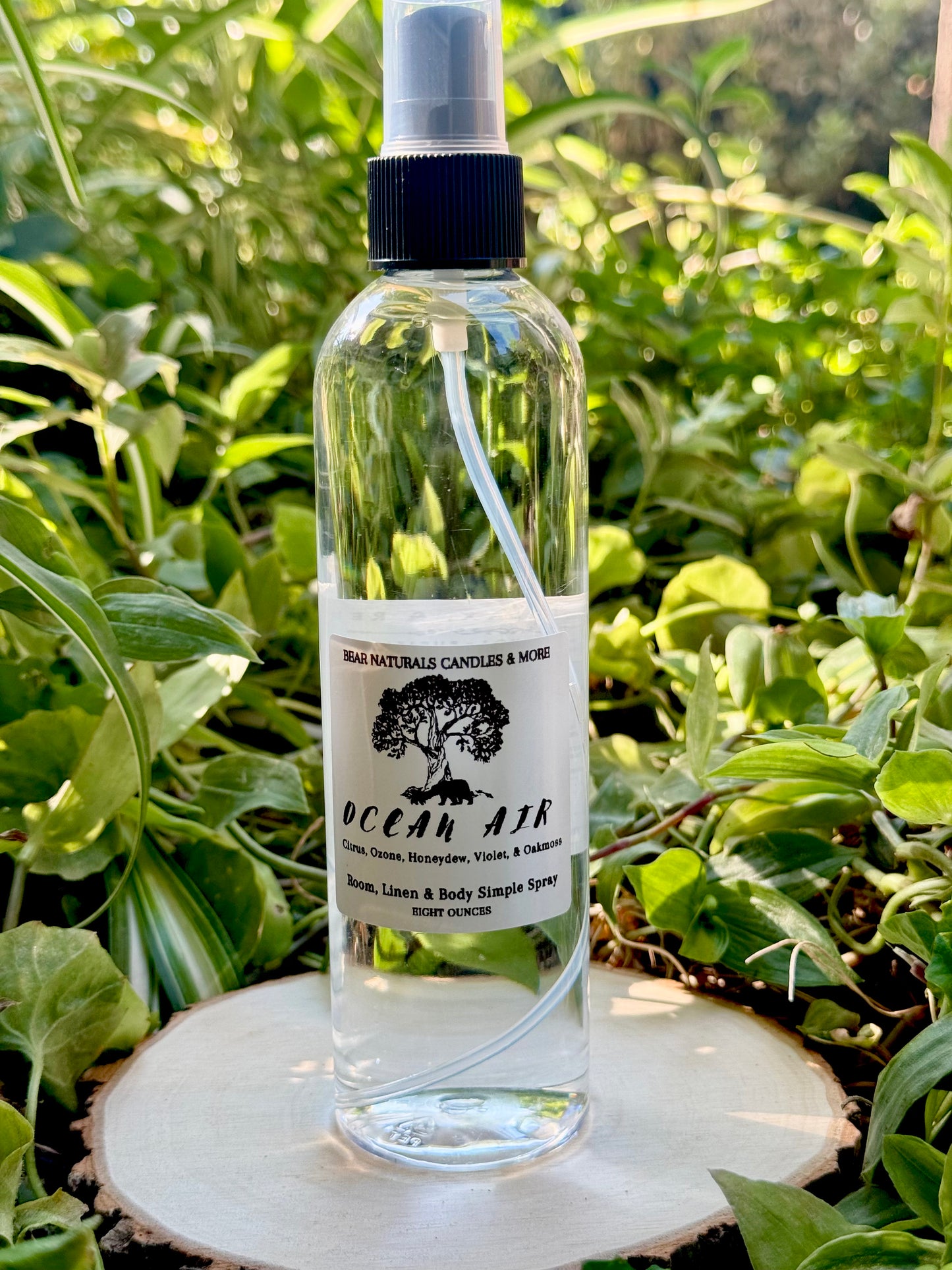 A clear eight ounce finger-tip spray bottle with black and white label on a wooden surface with green leaves in the background. The bottle contains the scent "Ocean Air".