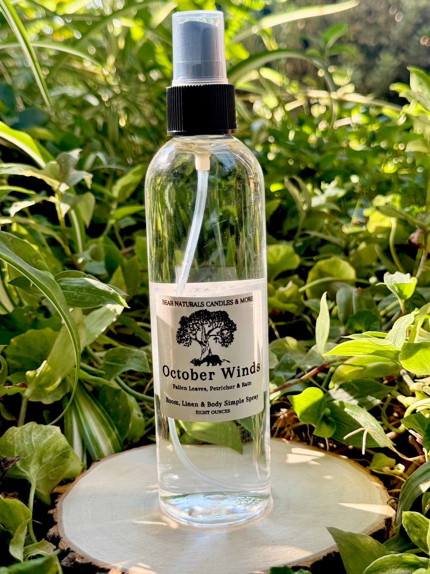 A clear eight ounce finger-tip spray bottle with black and white label on a wooden surface with green leaves in the background. The bottle contains the scent "October Winds"