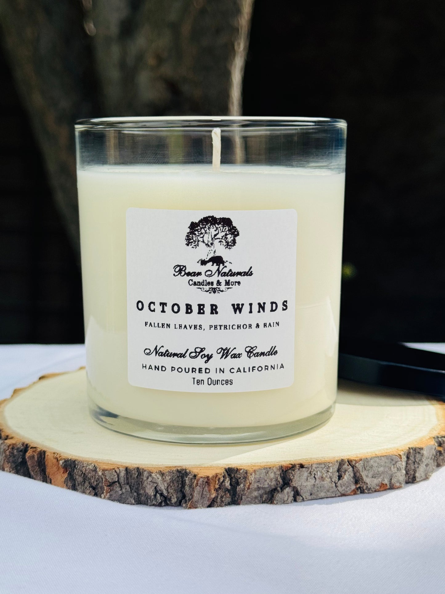 A white ten ounce candle in a clear glass jar with a black metal lid and white label with black writing sitting on a wooden surface. The candle is called "October Winds".